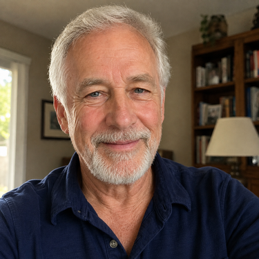 A close-up portrait of a smiling older man with gray hair and a beard, wearing a dark navy blue button-up shirt, set against a softly blurred indoor background with a bookshelf and lamp.